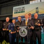 A group of eight officials, including maritime, fire, and police personnel, pose on stage at the State of the Port 2026 event at the Port of Los Angeles. Five individuals hold commemorative gold awards while standing around a central podium featuring the Port's logo