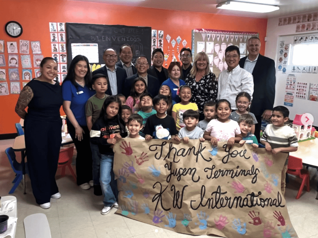 A group of smiling children and adults stand together in a brightly colored classroom at the Wilmington YMCA. The children in the foreground are holding a large, handmade brown paper banner that says "Thank You Yusen Terminals & KW International" decorated with colorful handprints. The adults, representing YTI and KW International, stand behind the children against an orange wall filled with student artwork and a chalkboard that says "Bienvenidos."