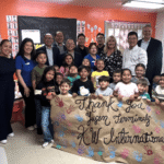 A group of smiling children and adults stand together in a brightly colored classroom at the Wilmington YMCA. The children in the foreground are holding a large, handmade brown paper banner that says "Thank You Yusen Terminals & KW International" decorated with colorful handprints. The adults, representing YTI and KW International, stand behind the children against an orange wall filled with student artwork and a chalkboard that says "Bienvenidos."
