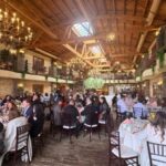A wide-angle view of a large, elegant banquet hall with high vaulted wooden ceilings and ornate chandeliers. Numerous guests are seated at round tables covered in white linens, enjoying a meal and conversation. The room features rustic brick walls, second-story indoor balconies decorated with greenery and red flowers, and a large hanging floral arrangement in the center of the space.