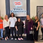 Un grupo de nueve profesionales posa para una foto en el vestíbulo de Yusen Terminals. Se encuentran frente a una gran pared iluminada con el logotipo de YTI. A la izquierda, una pantalla digital muestra el mensaje «Yusen Terminals da la bienvenida», seguido de varios logotipos de empresas de transporte. El grupo está formado por personal de la terminal, equipado con material de seguridad, y socios visitantes, vestidos con ropa informal de negocios.