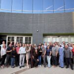 Un numeroso grupo de profesionales de diversos ámbitos del equipo de YTI posa al aire libre para una foto de grupo frente a un moderno edificio de ladrillo gris. Algunos miembros del equipo sostienen premios y un maletín plateado, mientras que otros sonríen y posan. El ambiente es festivo y de colaboración mientras se reúnen en un patio pavimentado bajo un cielo despejado.