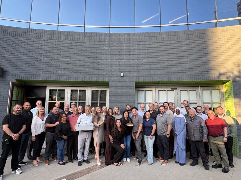 A large group of diverse professionals from the YTI team stands together outdoors for a group photo in front of a modern gray brick building. Some team members are holding awards and a silver briefcase, while others are smiling and posing. The atmosphere is celebratory and collaborative as they gather on a paved patio under a clear sky.