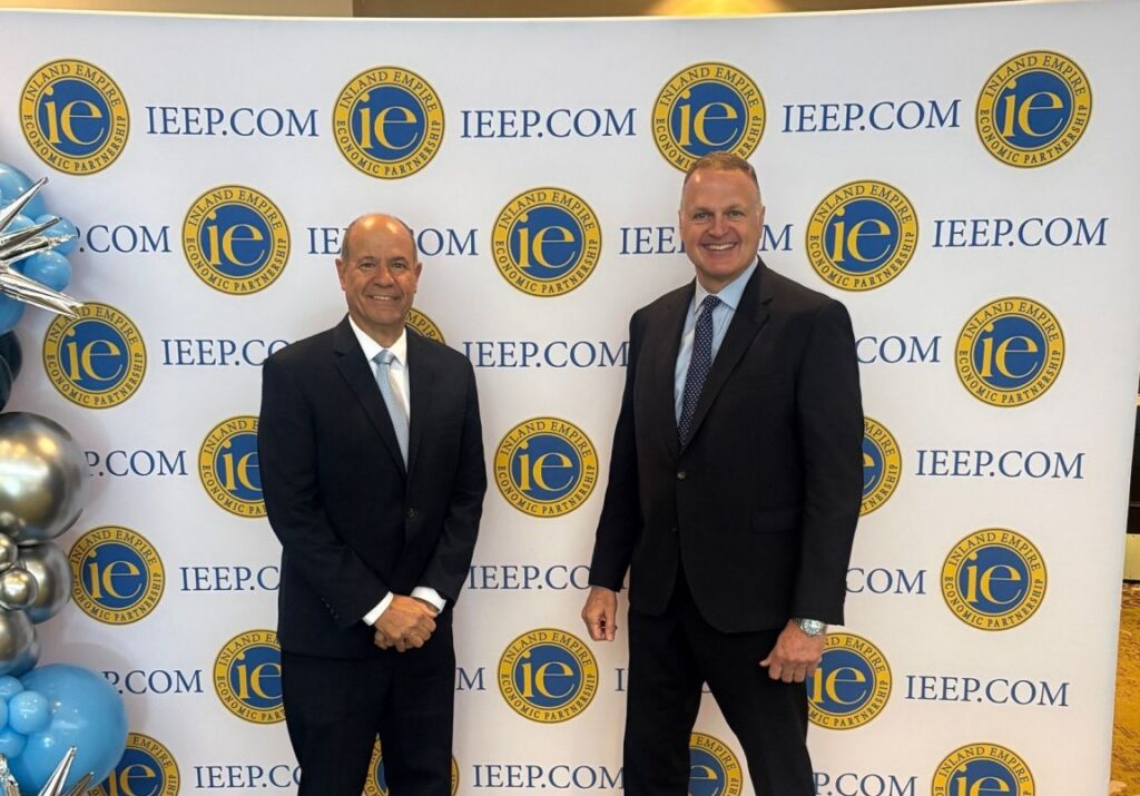 Alan McCorkle, President & CEO with Paul Granillo moderating for IEEP. Two business professionals posing at a logistics summit in front of a branded IEEP backdrop. To the left, a decorative arrangement of blue and silver balloons is visible. The scene is set on a patterned green and gold carpet at an indoor event venue.