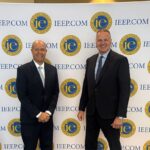 Alan McCorkle, presidente y director ejecutivo, con Paul Granillo como moderador en nombre del IEEP. Dos profesionales del mundo empresarial posan en una cumbre sobre logística frente a un telón de fondo con el logotipo del IEEP. A la izquierda se aprecia un arreglo decorativo de globos azules y plateados. La escena se desarrolla sobre una alfombra estampada en tonos verdes y dorados en un recinto para eventos cubierto.