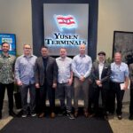 A group portrait at Yusen Terminals featuring eight men in business-casual and professional attire. The background includes the Yusen Terminals logo and a digital welcome sign listing several California port partners. The group is standing on a dark rug in a modern office lobby with indoor plants and a framed map on the wall Regional Maritime Cybersecurity Meeting