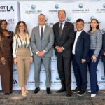 A professional group of ten individuals stands in a row, smiling for a group portrait at an event. They are positioned in front of a white step-and-repeat backdrop featuring repeating logos for "The Port of Los Angeles," "Port of Long Beach," and the "Los Angeles Area Chamber of Commerce."
