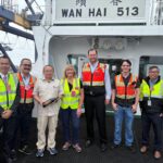 A group of seven people poses for a photo on the deck of a large ship, positioned beneath a white structural wall that features the text "WAN HAI 513" in bold black lettering, with traditional characters above it